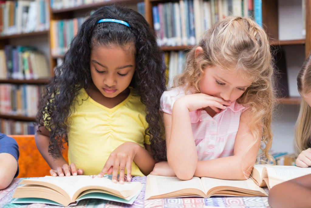 Approximately five year old African American girl reading with approximately five year old Caucasian girl with blonde hair in library.